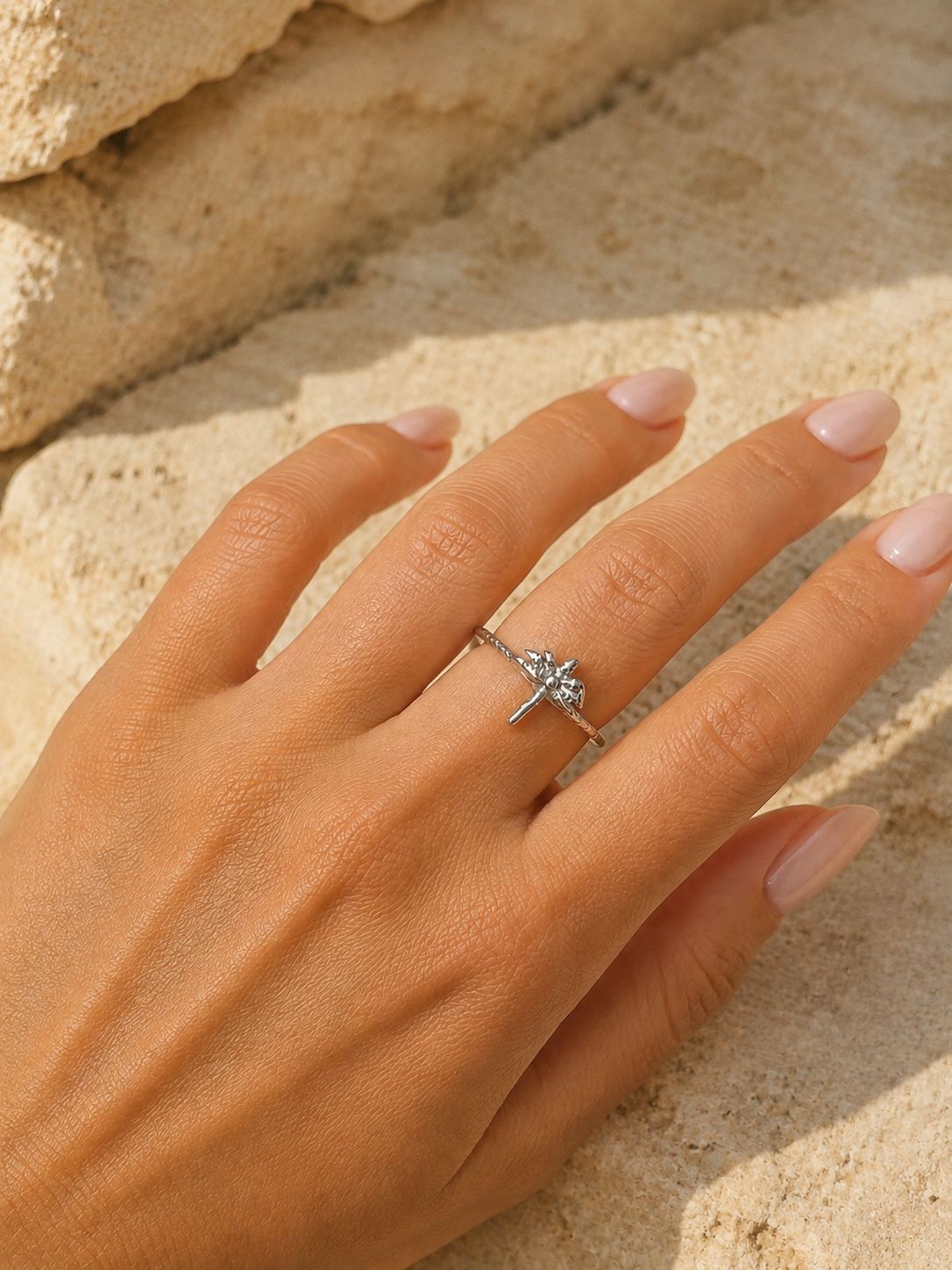 A silver ring with a small dragonfly design set with clear stones, worn on a hand with pale pink manicured nails, resting on a sandy beige background.