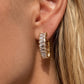 A close-up of a model's ear wearing a gold huggie hoop earring set with clear baguette-cut stones.