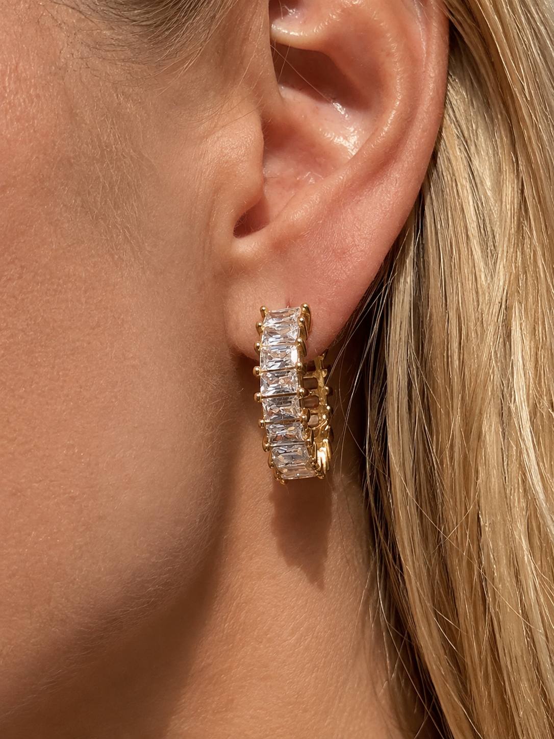 A close-up of a model's ear wearing a gold huggie hoop earring set with clear baguette-cut stones.