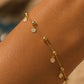 A delicate gold chain bracelet with small disc charms and bead details, shown worn on a person's wrist against a warm, softly blurred background.