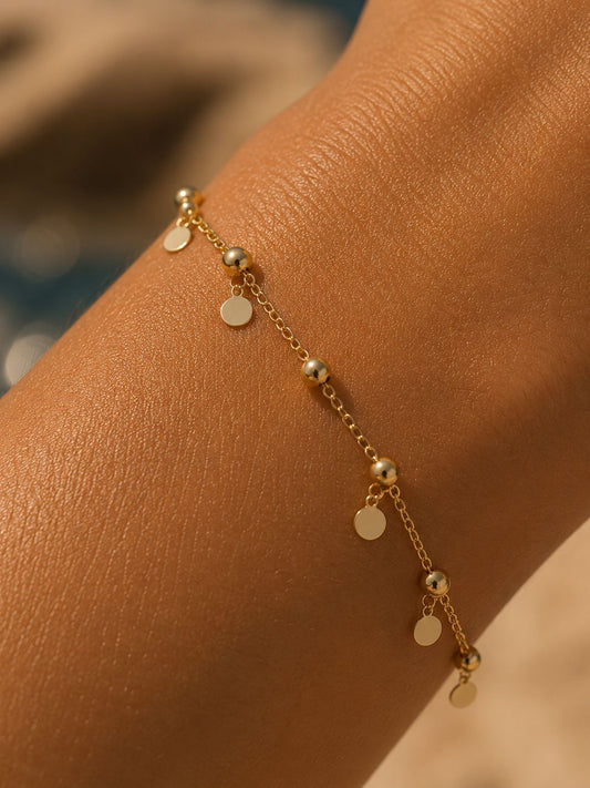 A delicate gold chain bracelet with small disc charms and bead details, shown worn on a person's wrist against a warm, softly blurred background.