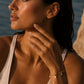 A close-up lifestyle shot of a woman outdoors near the water, wearing a delicate gold chain bracelet with small round charms on her wrist.