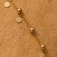 A gold chain bracelet with spherical beads and flat round disc charms, laid diagonally on a textured tan surface.