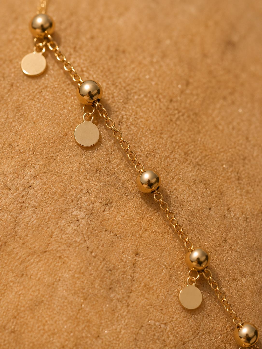 A gold chain bracelet with spherical beads and flat round disc charms, laid diagonally on a textured tan surface.
