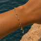 A delicate gold chain anklet with small disc charms is shown worn on a person's ankle, with a blurred background of blue water and a rock.