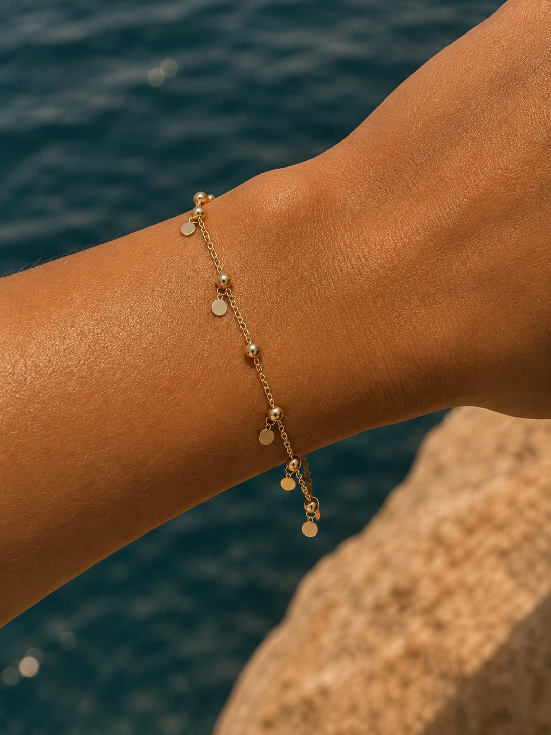 A delicate gold chain anklet with small disc charms is shown worn on a person's ankle, with a blurred background of blue water and a rock.