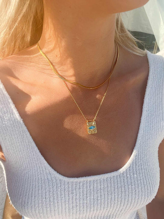 A woman wearing a delicate gold evil eye necklace with a square pendant featuring a blue zircon aquamarine gemstone and white zirconia gemstones, layered over a thin gold chain. Protection Jewelry.