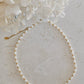 A delicate necklace made of small white pearls separated by tiny gold beads, arranged in an oval on a light textured surface with white flowers in the upper corner.