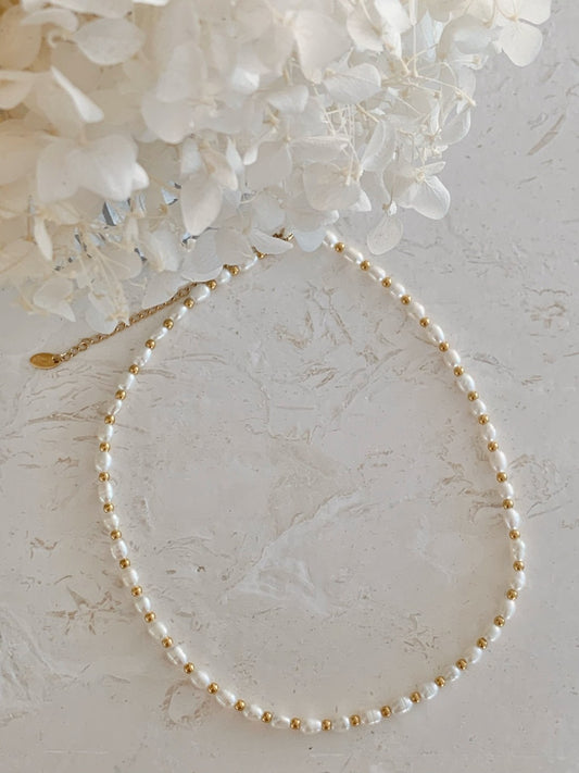 A delicate necklace made of small white pearls separated by tiny gold beads, arranged in an oval on a light textured surface with white flowers in the upper corner.