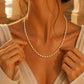 A delicate white pearl necklace, adjusting it with her hands. She is wearing a light beige linen top, and the background features warm, sunlit stone columns.