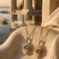 Gold and silver quartz crystal pendant necklaces displayed on textured sandstone beside still water and white lotus flowers.