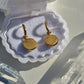 A pair of gold disc earrings resting in an open white jewelry box, with clear crystal points placed around them on a light surface.