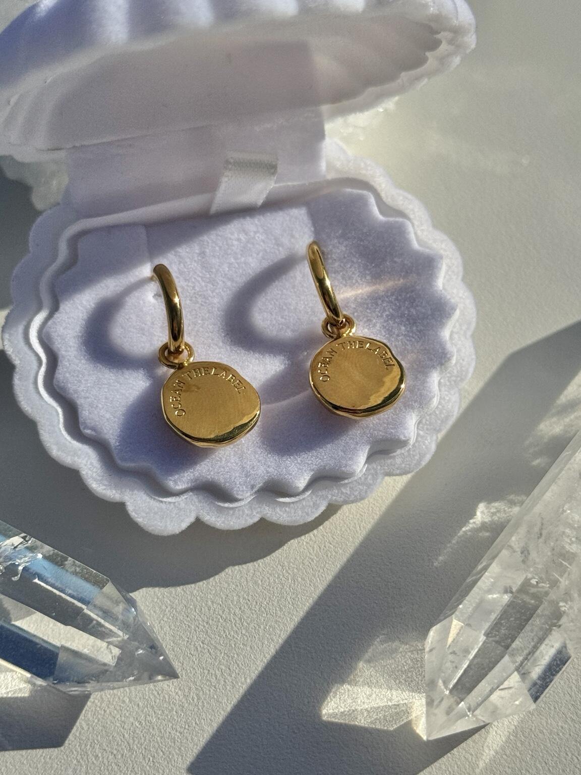 A pair of gold disc earrings resting in an open white jewelry box, with clear crystal points placed around them on a light surface.