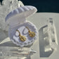 A pair of small gold drop earrings with textured circular charms, displayed inside an open white clamshell jewelry box on a white surface with clear crystal points around it.