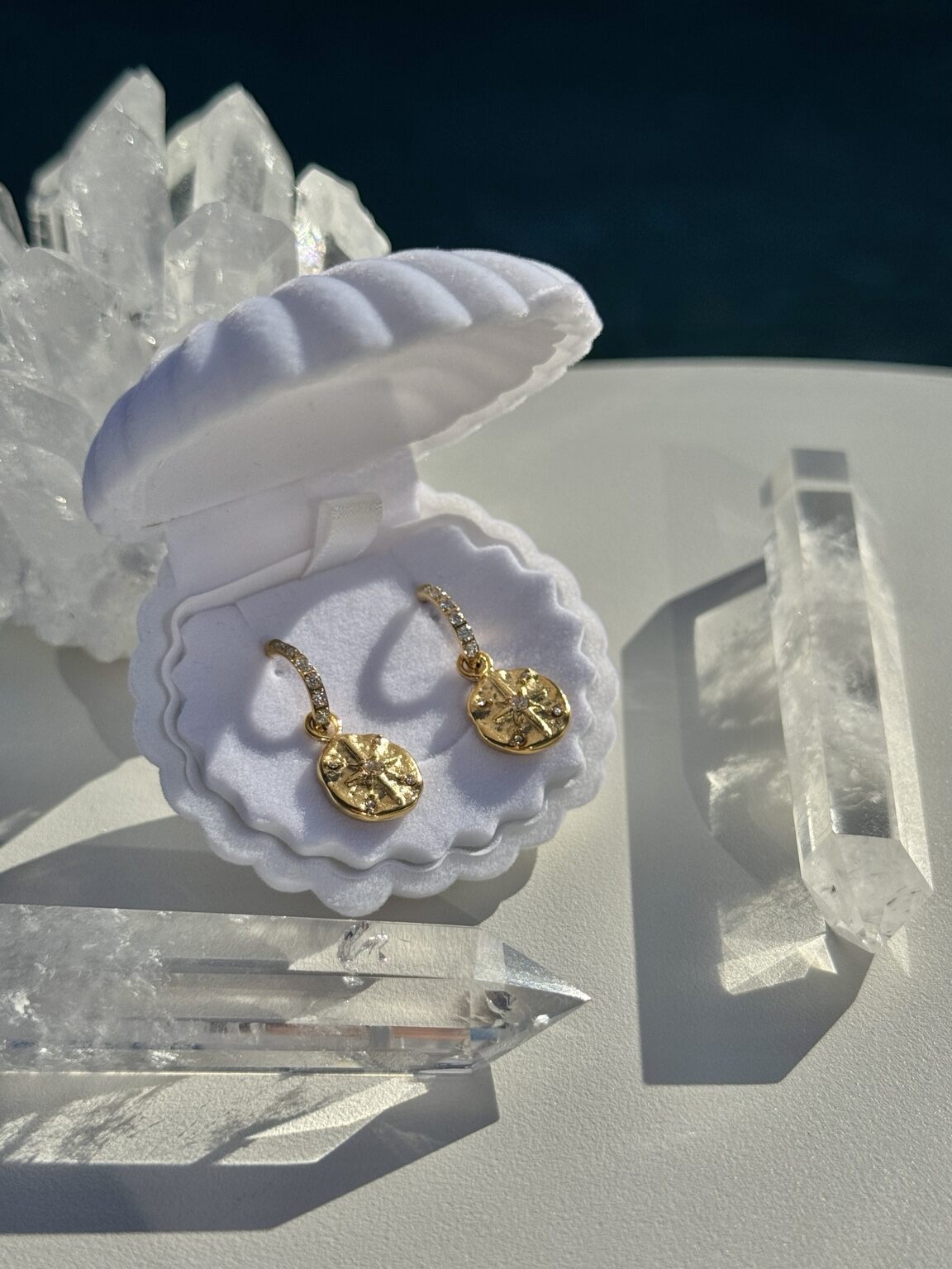 A pair of small gold drop earrings with textured circular charms, displayed inside an open white clamshell jewelry box on a white surface with clear crystal points around it.