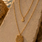 Two gold layered necklaces displayed on a beige stone outdoors near water at sunset. The shorter necklace has a small sunburst pendant, and the longer necklace has a round medallion pendant with an engraved sun design and a small clear stone center.