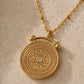A gold-toned circular pendant necklace with an embossed floral sunburst design and a small clear central stone, shown on a textured beige surface.