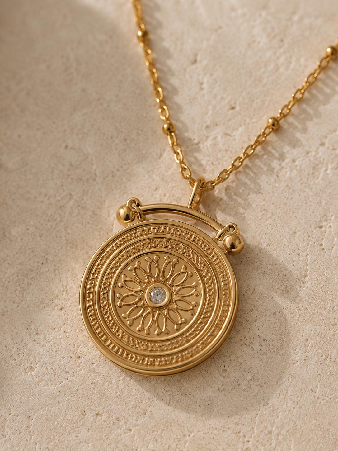 A gold-toned circular pendant necklace with an embossed floral sunburst design and a small clear central stone, shown on a textured beige surface.