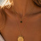 A close-up of a woman wearing a layered gold necklace set. The longer chain features a round, textured gold medallion pendant, while the shorter chain has a small dark green sunburst pendant.