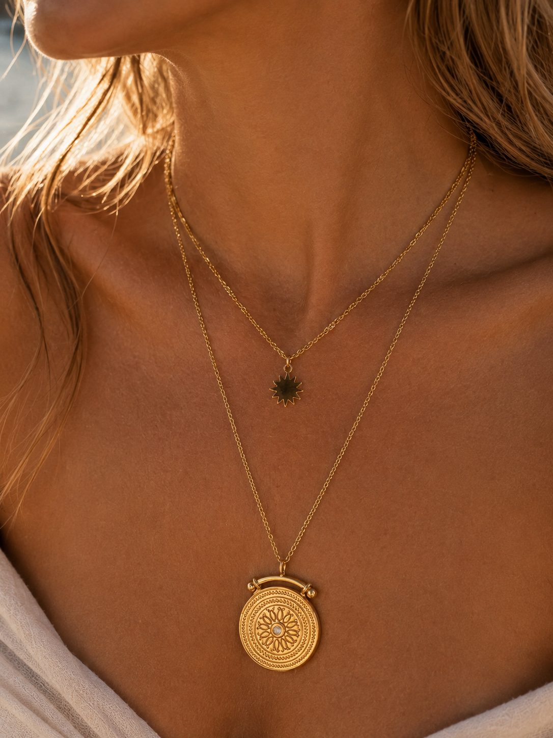 A close-up of a woman wearing a layered gold necklace set. The longer chain features a round, textured gold medallion pendant, while the shorter chain has a small dark green sunburst pendant.