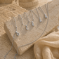 Five silver letter necklaces spelling 'OCEAN' are draped over a beige stone block in a coastal setting with sand and the sea in the background.