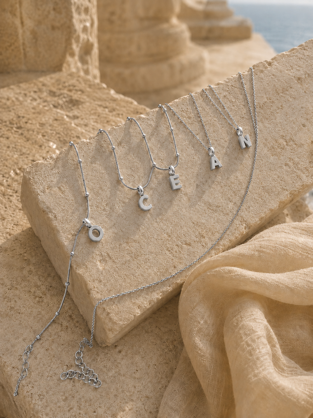 Five silver letter necklaces spelling 'OCEAN' are draped over a beige stone block in a coastal setting with sand and the sea in the background.
