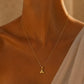 A close-up of a model's neck and upper chest wearing a delicate gold chain necklace with a small letter 'A' pendant, styled with a light beige draped top against a warm, softly lit background.