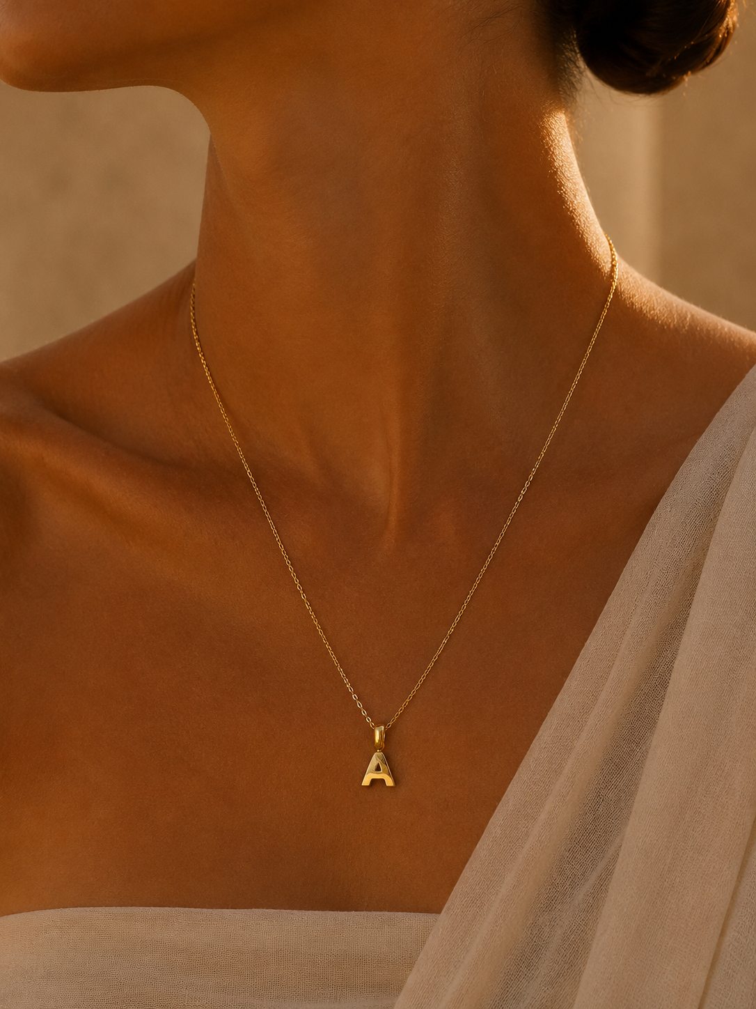 A close-up of a model's neck and upper chest wearing a delicate gold chain necklace with a small letter 'A' pendant, styled with a light beige draped top against a warm, softly lit background.