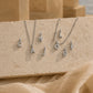 Four silver letter necklaces spelling out 'CAJOF' are displayed on a textured beige stone slab with draped beige fabric in the foreground and a softly blurred warm-toned architectural background.