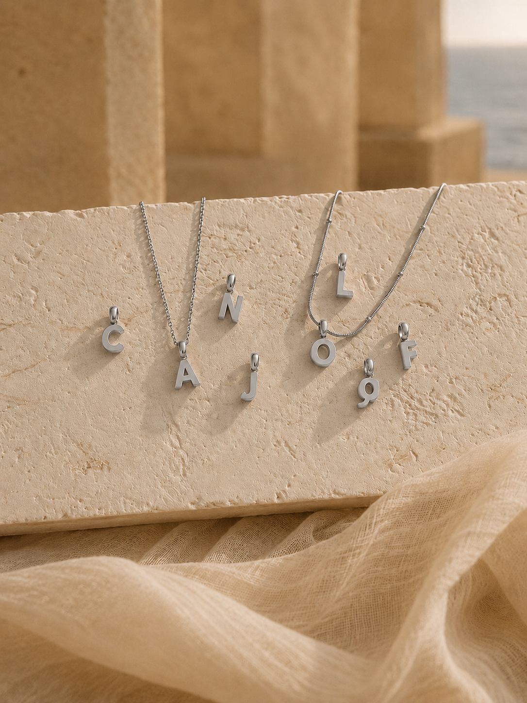 Four silver letter necklaces spelling out 'CAJOF' are displayed on a textured beige stone slab with draped beige fabric in the foreground and a softly blurred warm-toned architectural background.