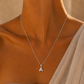 A close-up of a model's neck and upper chest wearing a delicate silver chain necklace with a small capital letter 'A' pendant, styled with a beige draped garment.