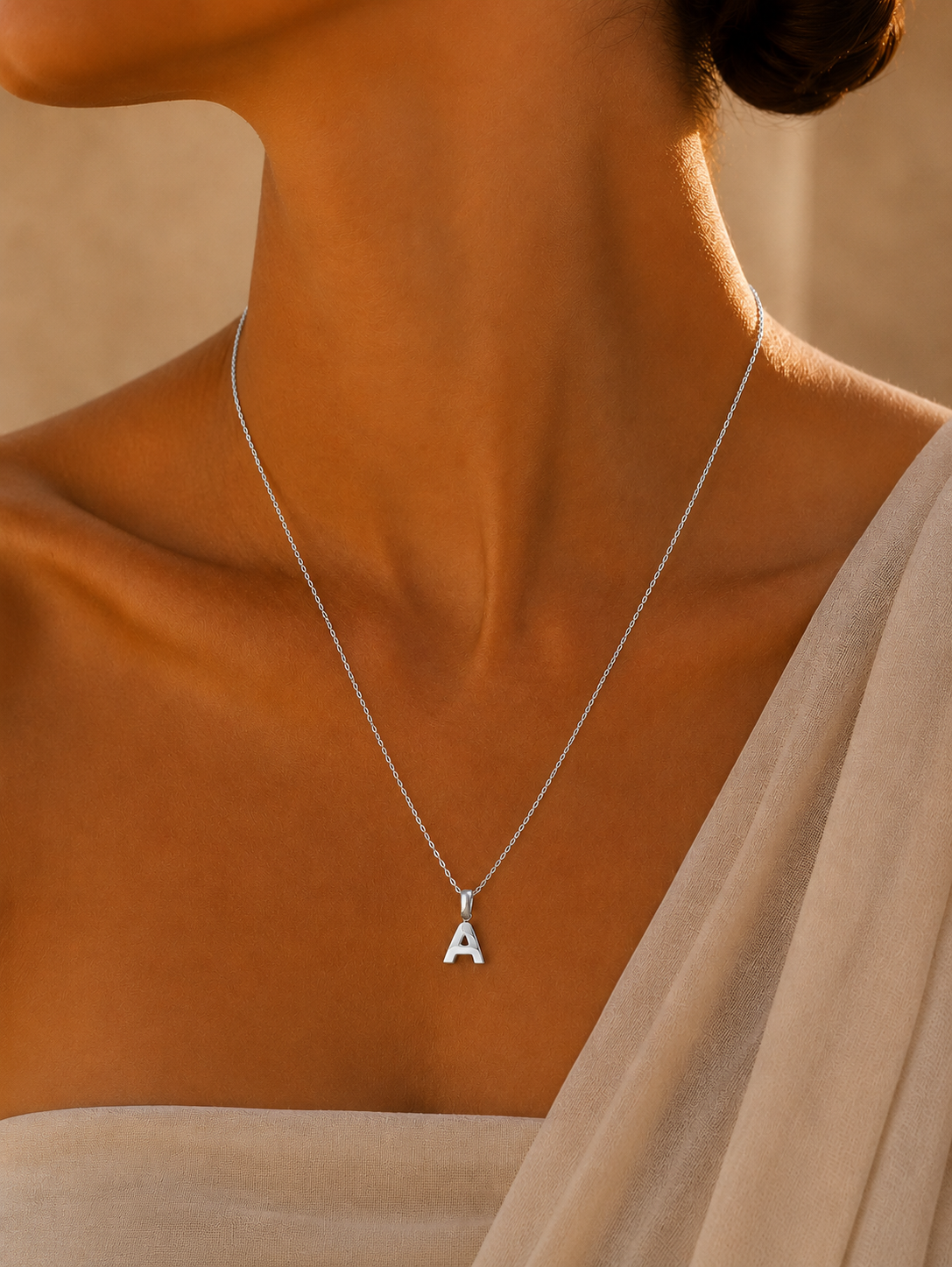 A close-up of a model's neck and upper chest wearing a delicate silver chain necklace with a small capital letter 'A' pendant, styled with a beige draped garment.