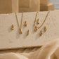 Gold letter necklaces with initial charms spelling 'CAJOF' and 'LÓN' arranged on a textured beige stone slab with draped fabric in the foreground.