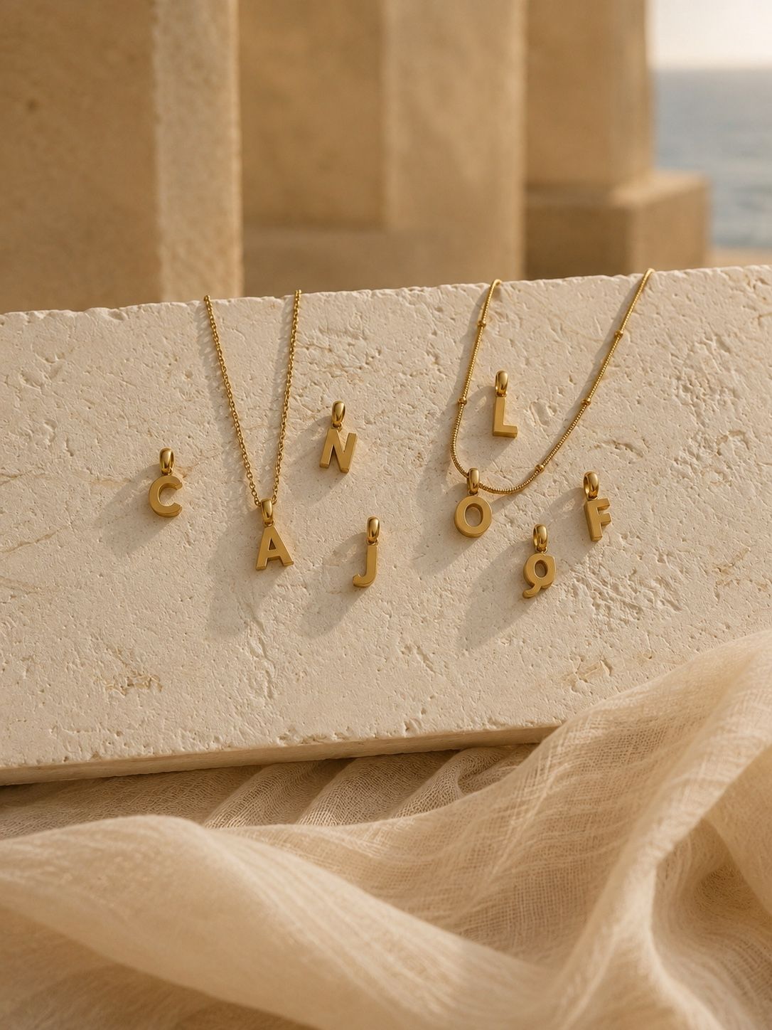 Gold letter necklaces with initial charms spelling 'CAJOF' and 'LÓN' arranged on a textured beige stone slab with draped fabric in the foreground.