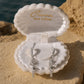 A pair of silver-toned drop earrings with clear stones, displayed in an open white shell-shaped jewelry box. The box lid is open and shows the brand name 'Ocean The Label' in gold foil.