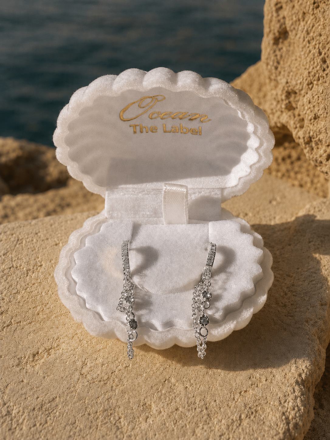 A pair of silver-toned drop earrings with clear stones, displayed in an open white shell-shaped jewelry box. The box lid is open and shows the brand name 'Ocean The Label' in gold foil.
