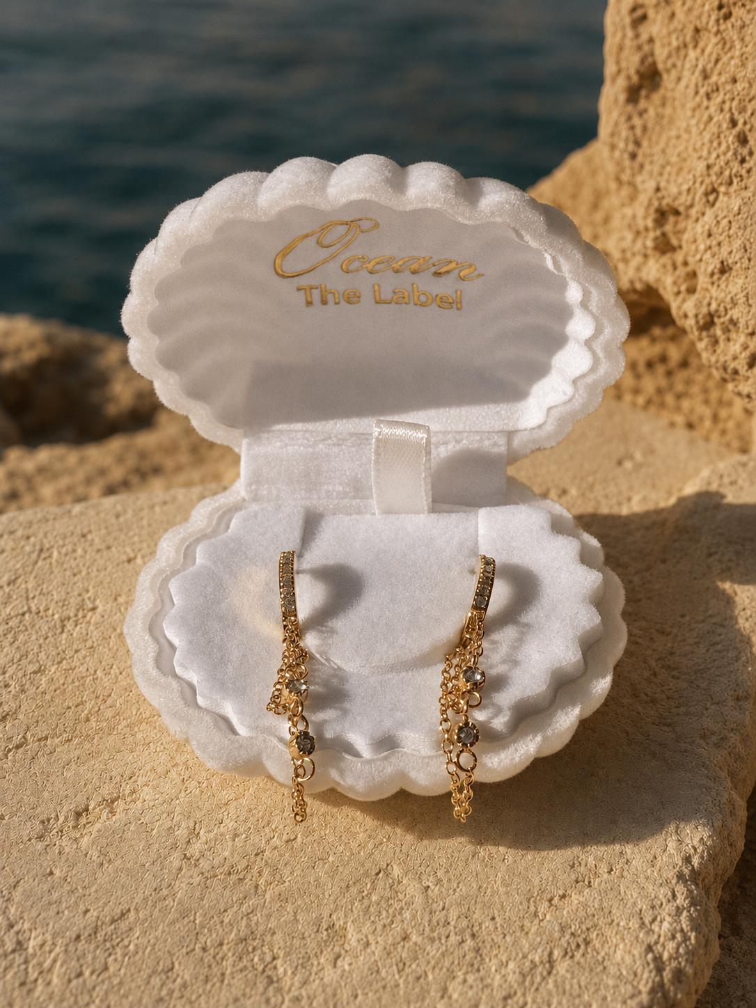 A pair of gold dangle earrings displayed in an open white shell-shaped jewelry box on a sandy surface with water in the background. The inside of the box lid is printed with the brand name 'Ocean The Label'.