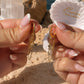 A close-up of two hands holding a pair of gold dangle earrings against a beach backdrop with sand, rocks, and ocean water. One earring has a small gold hoop with a dangling chain, and the other has a larger gold oval with a dangling chain.