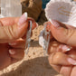 A close-up of two hands holding a pair of silver dangle earrings with clear stones, set against a beach background with sand, rocks, and a scalloped white jewelry box labeled 'Ocean The Label'.