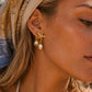 A close-up of a woman's ear wearing a gold starfish stud earring with a dangling white pearl, set against her hair and a patterned beige headscarf.