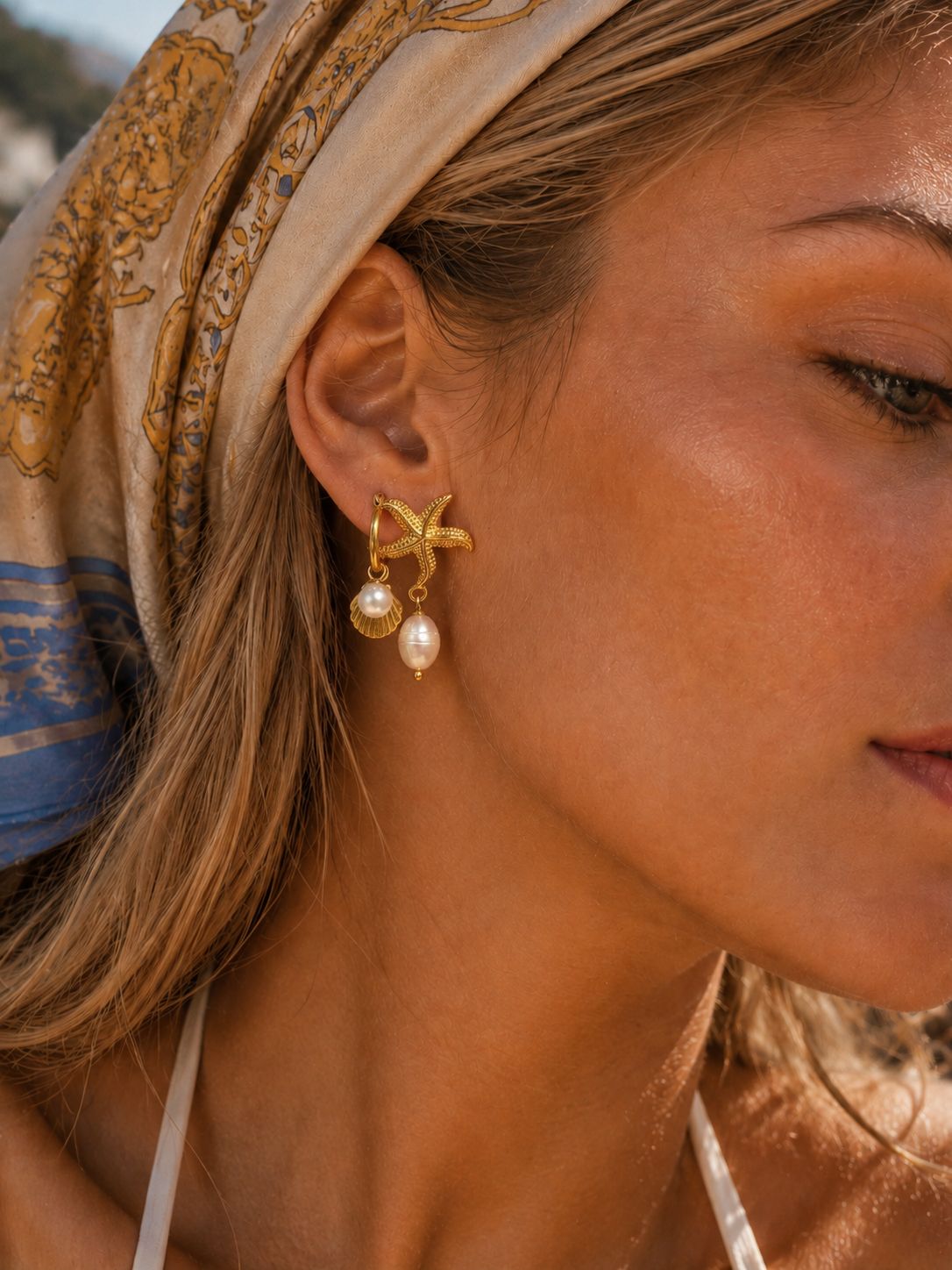 A close-up of a woman's ear wearing a gold starfish stud earring with a dangling white pearl, set against her hair and a patterned beige headscarf.