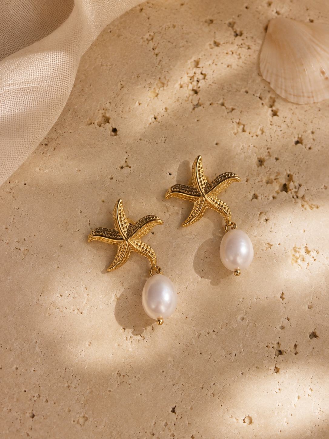 A pair of gold starfish drop earrings with white pearl pendants, styled on a sandy beige surface with soft fabric and a shell.