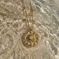 A gold pendant necklace with a circular sunburst design and a central white moonstone, resting on a wet stone surface in shallow water.