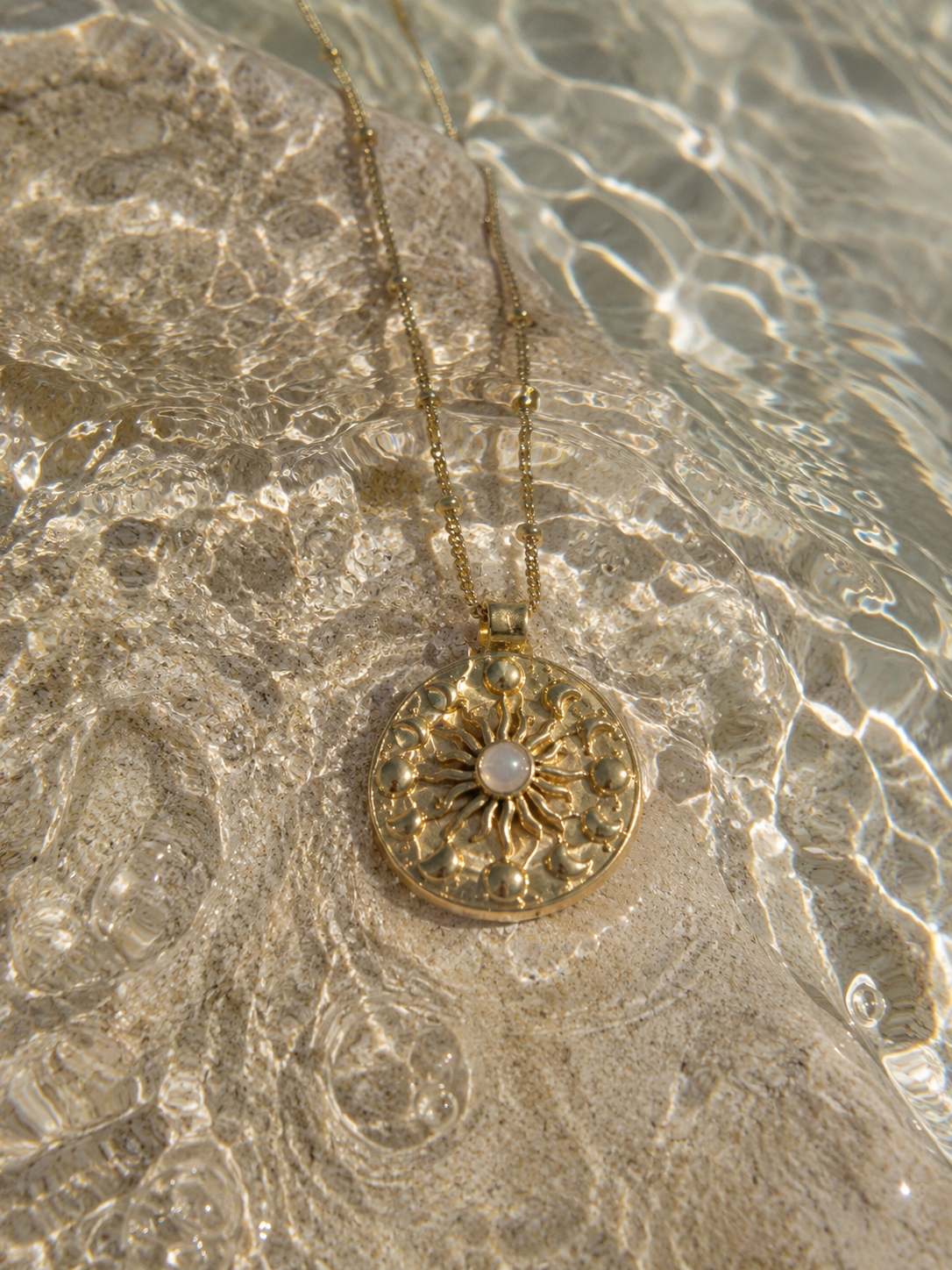 A gold pendant necklace with a circular sunburst design and a central white moonstone, resting on a wet stone surface in shallow water.