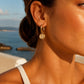 A
small gold hoop earring with a textured, diamond-like gold dangle against a warm, sunny outdoor background with the sea in the distance.