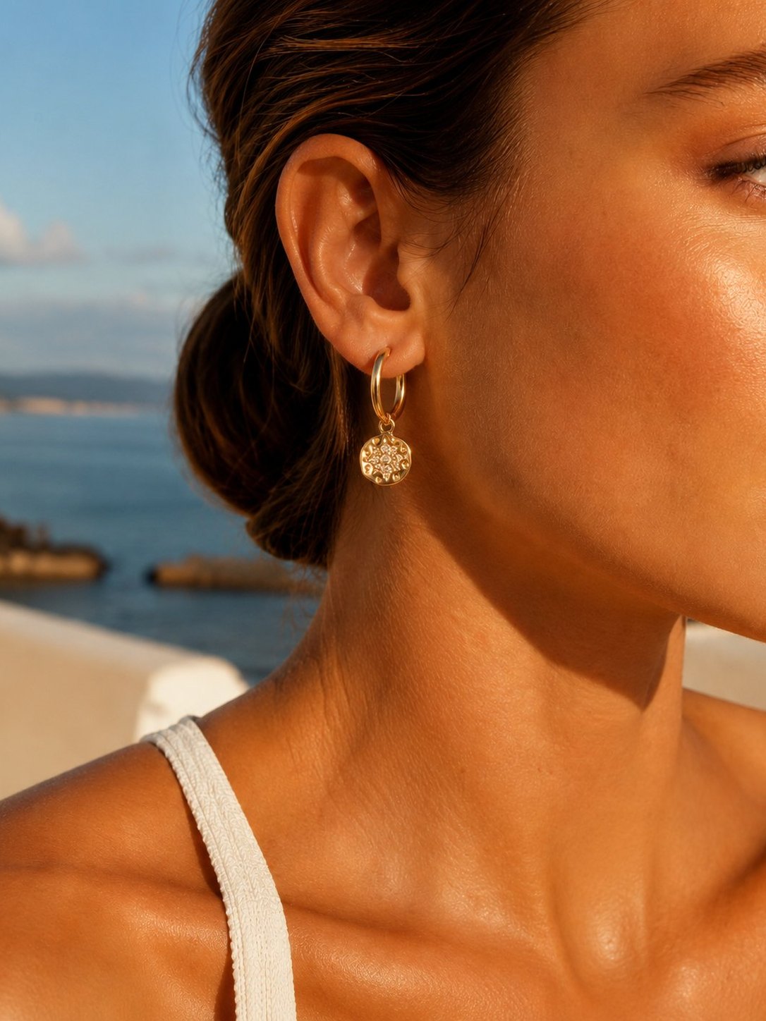 A
small gold hoop earring with a textured, diamond-like gold dangle against a warm, sunny outdoor background with the sea in the distance.