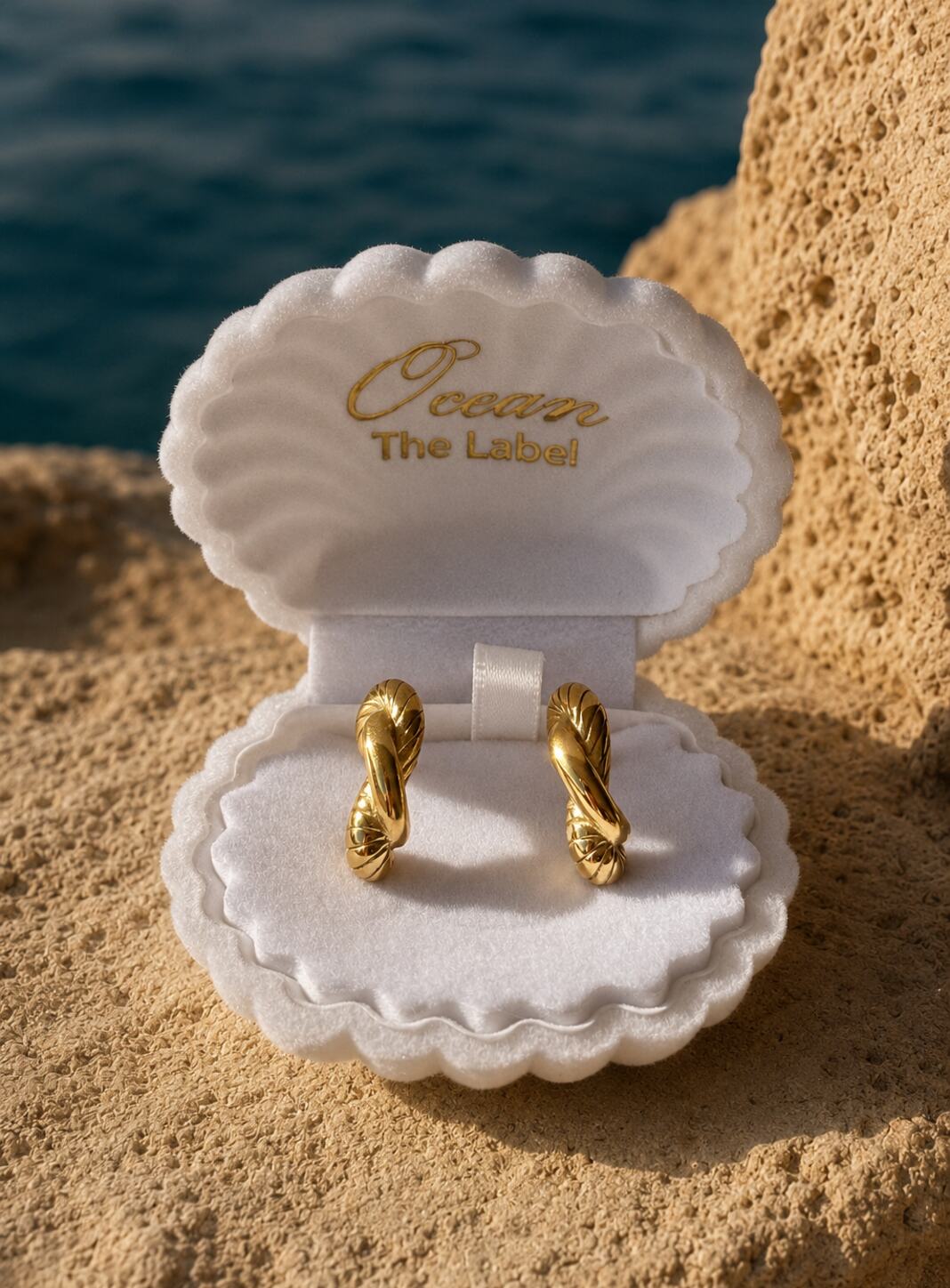 A pair of gold twisted hoop earrings displayed in an open white clamshell jewelry box on sandy beach ground with the ocean in the background. The inside of the shell is printed with the brand name 'Ocean The Label'.