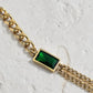 A gold chain bracelet featuring a rectangular green emerald-cut gemstone centerpiece, displayed on a textured white surface.