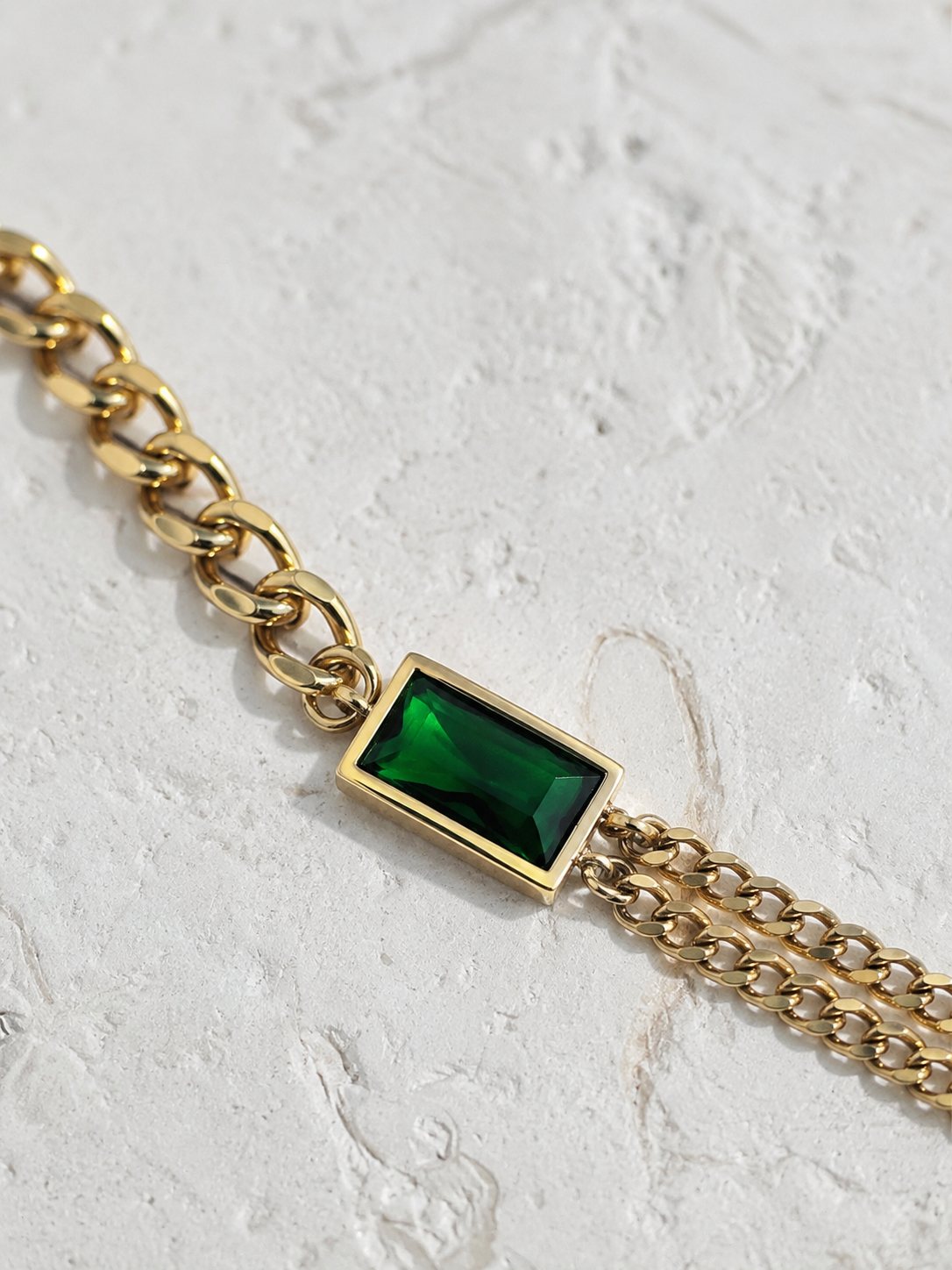 A gold chain bracelet featuring a rectangular green emerald-cut gemstone centerpiece, displayed on a textured white surface.