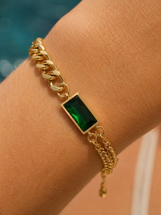 A gold chain bracelet with a rectangular green emerald-cut stone, worn on a person's wrist.